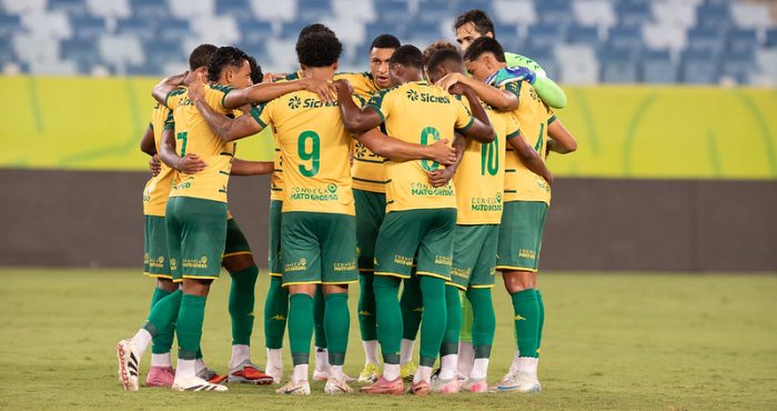 Foto do elenco do Cuiabá reunido em um círculo no centro do gramado. Os jogadores estão de costas, abraçados pelos ombros em um momento de união antes ou depois da partida. É possível ver os números 7, 9, 8 e 10 nos uniformes amarelos, além do goleiro à direita vestindo uniforme verde claro. Ao fundo, as arquibancadas do estádio estão vazias e iluminadas pelos refletores.