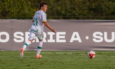 Jogador do Cuiabá Sub-20, vestindo uniforme branco com detalhes em verde e número 21, correndo próximo à linha lateral enquanto acompanha a bola em campo gramado.