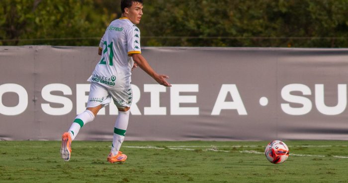 Jogador do Cuiabá Sub-20, vestindo uniforme branco com detalhes em verde e número 21, correndo próximo à linha lateral enquanto acompanha a bola em campo gramado.
