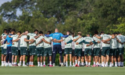 O elenco e a comissão técnica do Cuiabá formam um grande círculo (roda) no campo de treinamento. Os jogadores estão de costas, abraçados, enquanto um integrante da equipe técnica vestindo azul está no centro. O cenário mostra um gramado bem cuidado com árvores ao fundo sob a luz do dia.