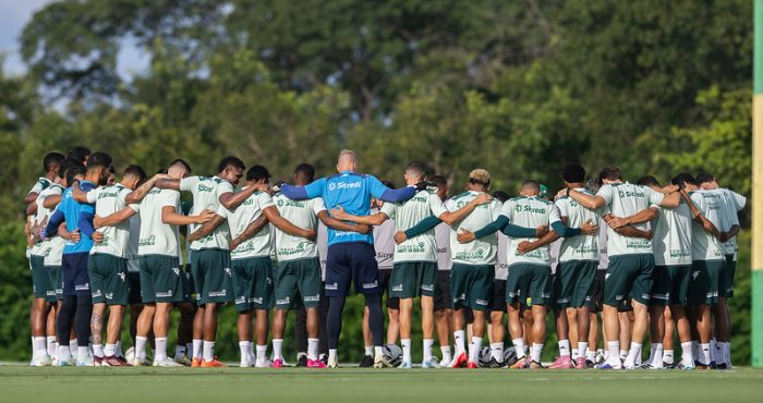 O elenco e a comissão técnica do Cuiabá formam um grande círculo (roda) no campo de treinamento. Os jogadores estão de costas, abraçados, enquanto um integrante da equipe técnica vestindo azul está no centro. O cenário mostra um gramado bem cuidado com árvores ao fundo sob a luz do dia.
