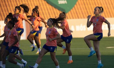 várias jogadoras da seleção Brasielria com seus uniformes de treino na cor pêssego, realizando exercícios de elevação de joelhos no CT Manoel Dresch. Os uniformes exibem o escudo da CBF (Confederação Brasileira de Futebol) e patrocinadores proeminentes como Itaú, Vivo e Guaraná Antarctica.