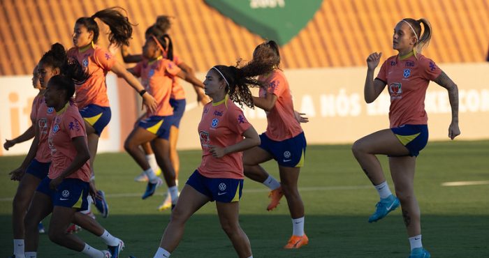 várias jogadoras da seleção Brasielria com seus uniformes de treino na cor pêssego, realizando exercícios de elevação de joelhos no CT Manoel Dresch. Os uniformes exibem o escudo da CBF (Confederação Brasileira de Futebol) e patrocinadores proeminentes como Itaú, Vivo e Guaraná Antarctica.