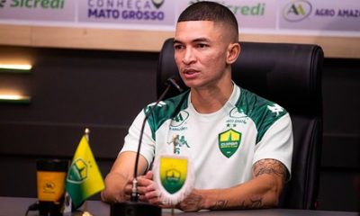 Lateral Marlon concede entrevista coletiva pelo Cuiabá, sentado à mesa com microfone e escudo do clube ao fundo.