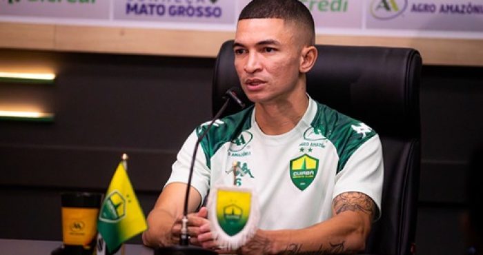 Lateral Marlon concede entrevista coletiva pelo Cuiabá, sentado à mesa com microfone e escudo do clube ao fundo.
