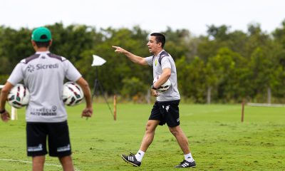 treinador Eduardo Barros com a camisa de treino cinza da Kappa, segurando uma bola e apontando para o campo, provavelmente orientando uma sessão tática.
