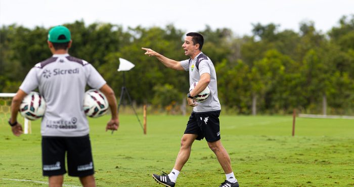 treinador Eduardo Barros com a camisa de treino cinza da Kappa, segurando uma bola e apontando para o campo, provavelmente orientando uma sessão tática.