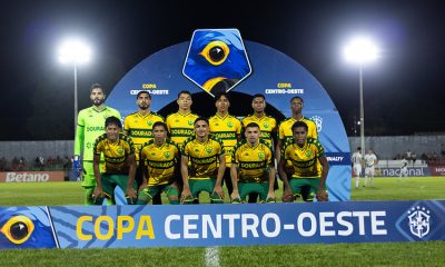 Time titular do Cuiabá posando para foto oficial antes da partida da Copa Centro-Oeste, com placa da competição ao fundo e estádio iluminado à noite.