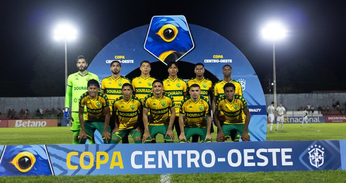 Time titular do Cuiabá posando para foto oficial antes da partida da Copa Centro-Oeste, com placa da competição ao fundo e estádio iluminado à noite.