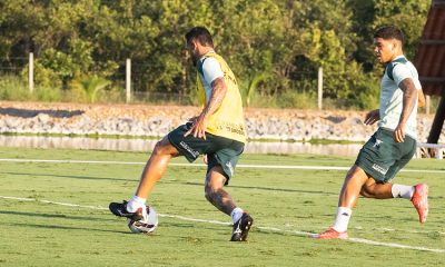 Dois jogadores de futebol profissionais durante um treino em um campo gramado ao ar livre, sob a luz do sol. O jogador em primeiro plano está de costas, vestindo um colete amarelo sobre o uniforme de treino verde-escuro e branco, controlando a bola com o pé direito. À direita, outro jogador corre em sua direção, acompanhando a jogada. Ambos usam shorts verdes com o patrocínio "Conheça Mato Grosso". Ao fundo, vê-se uma área arborizada e uma pequena faixa de água.