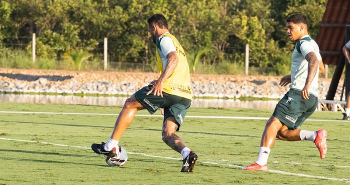 Dois jogadores de futebol profissionais durante um treino em um campo gramado ao ar livre, sob a luz do sol. O jogador em primeiro plano está de costas, vestindo um colete amarelo sobre o uniforme de treino verde-escuro e branco, controlando a bola com o pé direito. À direita, outro jogador corre em sua direção, acompanhando a jogada. Ambos usam shorts verdes com o patrocínio "Conheça Mato Grosso". Ao fundo, vê-se uma área arborizada e uma pequena faixa de água.