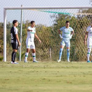 “Jogadores das categorias de base do Cuiabá EC posicionados na área durante partida contra o Corinthians em campo de treinamento, com uniforme branco do Dourado e rede do gol ao fundo.”