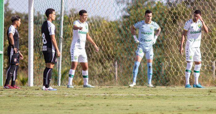 “Jogadores das categorias de base do Cuiabá EC posicionados na área durante partida contra o Corinthians em campo de treinamento, com uniforme branco do Dourado e rede do gol ao fundo.”