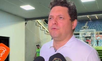 Entrevista em zona mista de estádio de futebol, com homem de camisa polo branca falando à imprensa. Microfones de emissoras de TV e escudo de clube de futebol aparecem em destaque, arquibancadas ao fundo.