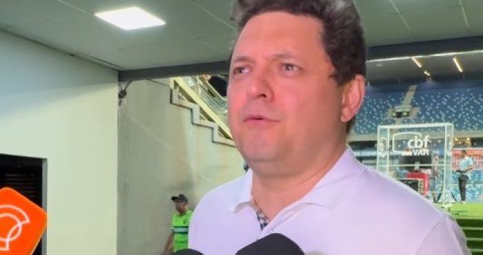 Entrevista em zona mista de estádio de futebol, com homem de camisa polo branca falando à imprensa. Microfones de emissoras de TV e escudo de clube de futebol aparecem em destaque, arquibancadas ao fundo.