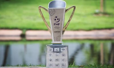 Troféu oficial do Campeonato Mato-Grossense da FMF exibido ao ar livre, com design moderno em tons prateados e dourados, apoiado sobre base decorada com ícones representando elementos do estado de Mato Grosso.