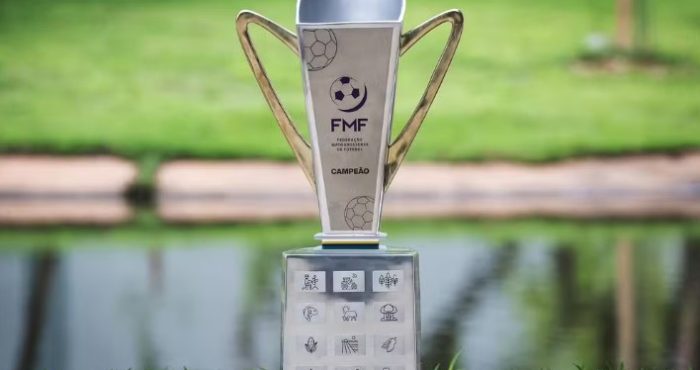 Troféu oficial do Campeonato Mato-Grossense da FMF exibido ao ar livre, com design moderno em tons prateados e dourados, apoiado sobre base decorada com ícones representando elementos do estado de Mato Grosso.