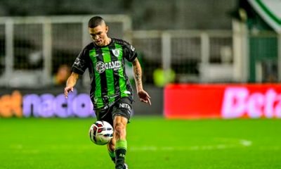 Fotografia de ação do jogador Marlon concentrado durante uma partida noturna. Ele está em movimento, preparando-se para dominar ou chutar a bola de futebol que está no ar à sua frente. Veste o uniforme listrado em verde e preto. O fundo mostra as luzes do estádio e placas de publicidade desfocadas.