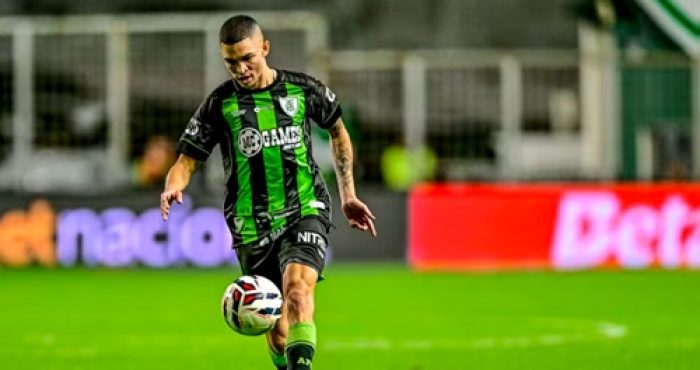 Fotografia de ação do jogador Marlon concentrado durante uma partida noturna. Ele está em movimento, preparando-se para dominar ou chutar a bola de futebol que está no ar à sua frente. Veste o uniforme listrado em verde e preto. O fundo mostra as luzes do estádio e placas de publicidade desfocadas.