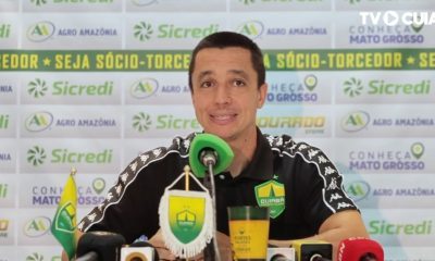Fotografia de Eduardo Barros, integrante da comissão técnica do Cuiabá Esporte Clube, durante uma entrevista. Ele veste uma camisa polo preta do time e está à frente de um painel com logotipos de patrocinadores como Sicredi e Agro Amazônia.