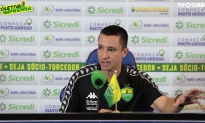 Eduardo Barros concede entrevista coletiva após jogo do Cuiabá, sentado à mesa com microfone e bandeira do clube ao fundo com patrocinadores.