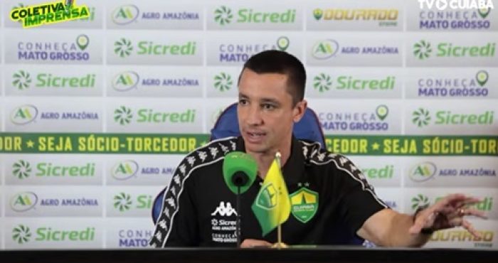 Eduardo Barros concede entrevista coletiva após jogo do Cuiabá, sentado à mesa com microfone e bandeira do clube ao fundo com patrocinadores.