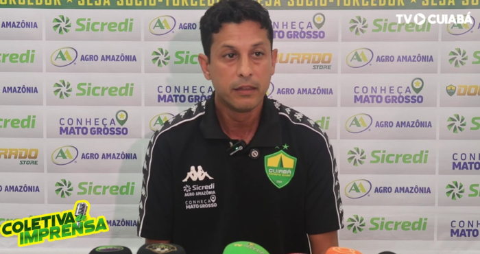 auxiliar técnico Gustavo Almeida concede entrevista coletiva após jogo do Cuiabá, sentado à mesa com microfone e bandeira do clube ao fundo com patrocinadores.