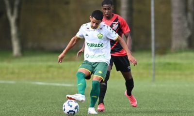 Jogador do Cuiabá Sub-20, vestindo uniforme branco e verde, conduz a bola enquanto é marcado por atleta do Athletico Paranaense, que usa uniforme vermelho e preto, durante partida do Campeonato Brasileiro Sub-20 em campo de grama sintética.