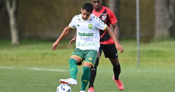 Jogador do Cuiabá Sub-20, vestindo uniforme branco e verde, conduz a bola enquanto é marcado por atleta do Athletico Paranaense, que usa uniforme vermelho e preto, durante partida do Campeonato Brasileiro Sub-20 em campo de grama sintética.