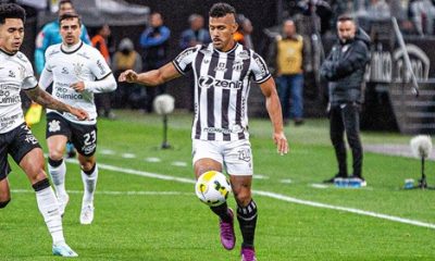 O jogador Fernando Sobral, vestindo o uniforme listrado em preto e branco do Ceará SC, controla a bola com o pé direito durante uma partida em um gramado molhado. Ele está cercado por dois jogadores do Corinthians, que vestem uniformes brancos. Ao fundo, vê-se a área técnica com membros da comissão e arquibancadas desfocadas.