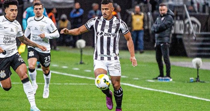 O jogador Fernando Sobral, vestindo o uniforme listrado em preto e branco do Ceará SC, controla a bola com o pé direito durante uma partida em um gramado molhado. Ele está cercado por dois jogadores do Corinthians, que vestem uniformes brancos. Ao fundo, vê-se a área técnica com membros da comissão e arquibancadas desfocadas.