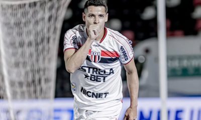Fotografia de meio corpo do jogador de futebol Matheus Barbosa comemorando um gol pelo Botafogo-SP. Ele veste o uniforme branco com listras horizontais vermelhas e pretas no peito e o patrocínio "Estrela Bet". O jogador está com o punho fechado próximo à boca, em um gesto de silêncio ou beijo, olhando diretamente para a câmera. Ao fundo, vê-se a rede do gol e as arquibancadas desfocadas.