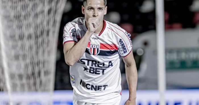 Fotografia de meio corpo do jogador de futebol Matheus Barbosa comemorando um gol pelo Botafogo-SP. Ele veste o uniforme branco com listras horizontais vermelhas e pretas no peito e o patrocínio "Estrela Bet". O jogador está com o punho fechado próximo à boca, em um gesto de silêncio ou beijo, olhando diretamente para a câmera. Ao fundo, vê-se a rede do gol e as arquibancadas desfocadas.
