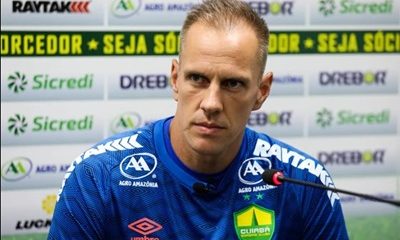 Fotografia de corpo inteiro do goleiro João Carlos em campo durante um jogo ou treino. Ele veste o uniforme azul de goleiro do Cuiabá, composto por camisa azul com detalhes vermelhos e o patrocínio "Drebor" e "Sicredi", além de calças térmicas e shorts azuis. Ele segura uma bola de futebol com a mão direita e mantém o braço esquerdo estendido, gesticulando para orientar o time. Ao fundo, veem-se as arquibancadas vazias de um estádio e placas de publicidade.