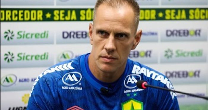 Fotografia de corpo inteiro do goleiro João Carlos em campo durante um jogo ou treino. Ele veste o uniforme azul de goleiro do Cuiabá, composto por camisa azul com detalhes vermelhos e o patrocínio "Drebor" e "Sicredi", além de calças térmicas e shorts azuis. Ele segura uma bola de futebol com a mão direita e mantém o braço esquerdo estendido, gesticulando para orientar o time. Ao fundo, veem-se as arquibancadas vazias de um estádio e placas de publicidade.