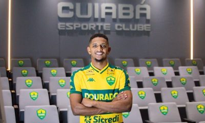 Fotografia de plano médio do atacante Hernandes, agora com os braços cruzados e um sorriso confiante. Ele está no centro de uma sala de imprensa ou auditório. Ao fundo, as poltronas cinzas possuem o escudo do Cuiabá bordado nos encostos. No topo da parede cinza ao fundo, destaca-se o letreiro metálico iluminado: "CUIABÁ ESPORTE CLUBE". A iluminação é suave, com faixas de luz verticais nas laterais.