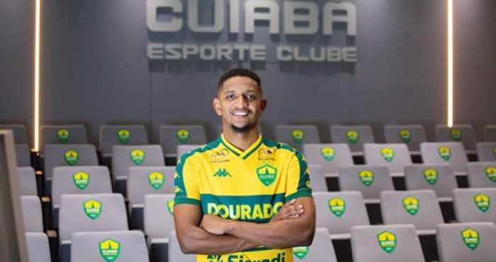Fotografia de plano médio do atacante Hernandes, agora com os braços cruzados e um sorriso confiante. Ele está no centro de uma sala de imprensa ou auditório. Ao fundo, as poltronas cinzas possuem o escudo do Cuiabá bordado nos encostos. No topo da parede cinza ao fundo, destaca-se o letreiro metálico iluminado: "CUIABÁ ESPORTE CLUBE". A iluminação é suave, com faixas de luz verticais nas laterais.