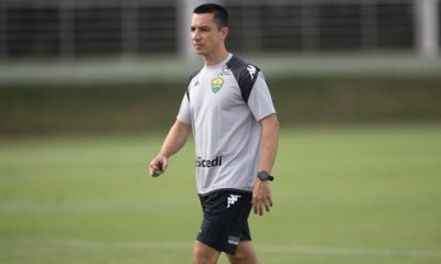 Fotografia em plano médio do técnico Eduardo Barros durante um treinamento no Cuiabá Esporte Clube. Ele é um homem de pele clara, cabelos curtos e escuros, e veste o uniforme de treino oficial: uma camiseta cinza com detalhes pretos nos ombros, o escudo do clube no peito e o patrocínio da Sicredi. Ele usa um relógio de pulso preto e caminha pelo gramado verde, que aparece levemente desfocado ao fundo.
