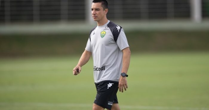 Fotografia em plano médio do técnico Eduardo Barros durante um treinamento no Cuiabá Esporte Clube. Ele é um homem de pele clara, cabelos curtos e escuros, e veste o uniforme de treino oficial: uma camiseta cinza com detalhes pretos nos ombros, o escudo do clube no peito e o patrocínio da Sicredi. Ele usa um relógio de pulso preto e caminha pelo gramado verde, que aparece levemente desfocado ao fundo.