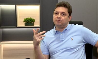 Cristiano dresch sentado em uma cadeira durante entrevista, vestindo camisa polo azul clara, gesticulando com a mão enquanto fala, com expressão séria e ambiente moderno ao fundo.