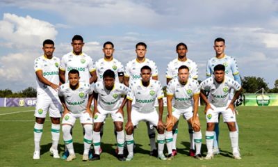 Foto oficial do time Sub-20 do Cuiabá Esporte Clube posado no gramado antes do jogo, com os atletas uniformizados de branco com detalhes verdes e amarelos. Ao fundo, céu azul com nuvens e a faixa do campeonato.