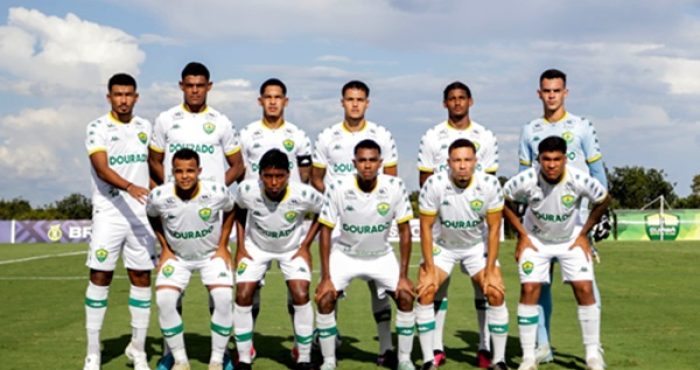 Foto oficial do time Sub-20 do Cuiabá Esporte Clube posado no gramado antes do jogo, com os atletas uniformizados de branco com detalhes verdes e amarelos. Ao fundo, céu azul com nuvens e a faixa do campeonato.