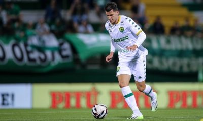 "Jogador do Cuiabá, usando uniforme branco com detalhes verdes, conduz a bola durante partida, com torcida e bandeiras verdes ao fundo."
