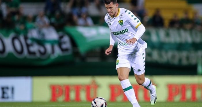 "Jogador do Cuiabá, usando uniforme branco com detalhes verdes, conduz a bola durante partida, com torcida e bandeiras verdes ao fundo."