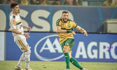 O Lateral Sander com a camisa amarela e verde do Cuiabá chuta a bola com o pé direito, em uma partida anrena pantanal. O marcador do novorizontino, vestindo camisa branca com detalhes pretos e amarelos e calção branco, observa a jogada ao lado. O campo é de grama, e a arquibancada azul e branca aparece desfocada ao fundo.