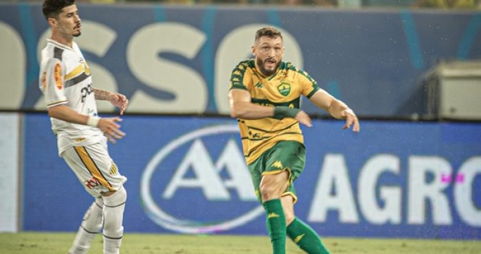 O Lateral Sander com a camisa amarela e verde do Cuiabá chuta a bola com o pé direito, em uma partida anrena pantanal. O marcador do novorizontino, vestindo camisa branca com detalhes pretos e amarelos e calção branco, observa a jogada ao lado. O campo é de grama, e a arquibancada azul e branca aparece desfocada ao fundo.