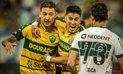 Dois jogadores do Cuiabá, em uniformes amarelos e verdes, se abraçam e comemoram um gol com expressões de alegria intensa. Um jogador adversário, em uniforme branco e verde com o número 73 nas costas, observa ao lado direito.