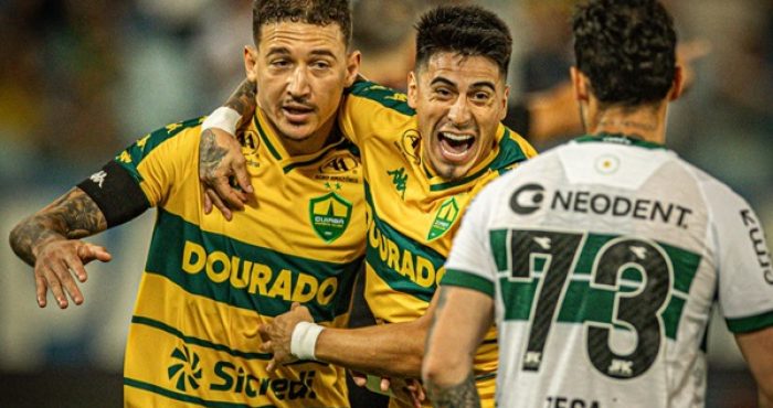 Dois jogadores do Cuiabá, em uniformes amarelos e verdes, se abraçam e comemoram um gol com expressões de alegria intensa. Um jogador adversário, em uniforme branco e verde com o número 73 nas costas, observa ao lado direito.