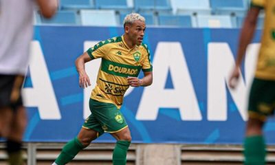 Foto de ação do atacante Juan Christian durante uma partida pelo Cuiabá. Ele aparece correndo no gramado, focado na jogada. Veste o uniforme do Cuiabá: camisa amarela com uma faixa horizontal verde (escrito "DOURADO") e calções verdes. Ao fundo, vê-se a arquibancada azul de um estádio e parte de uma placa publicitária. O jogador tem o cabelo curto e descolorido de loiro.