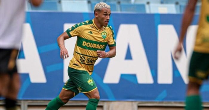 Foto de ação do atacante Juan Christian durante uma partida pelo Cuiabá. Ele aparece correndo no gramado, focado na jogada. Veste o uniforme do Cuiabá: camisa amarela com uma faixa horizontal verde (escrito "DOURADO") e calções verdes. Ao fundo, vê-se a arquibancada azul de um estádio e parte de uma placa publicitária. O jogador tem o cabelo curto e descolorido de loiro.